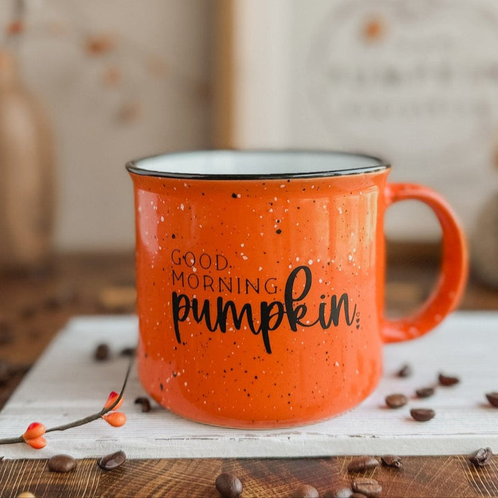 An orange ceramic coffee mug with black speckles and the text 'Good Morning Pumpkin' printed on it, placed on a table with a pumpkin-themed decoration in the background.