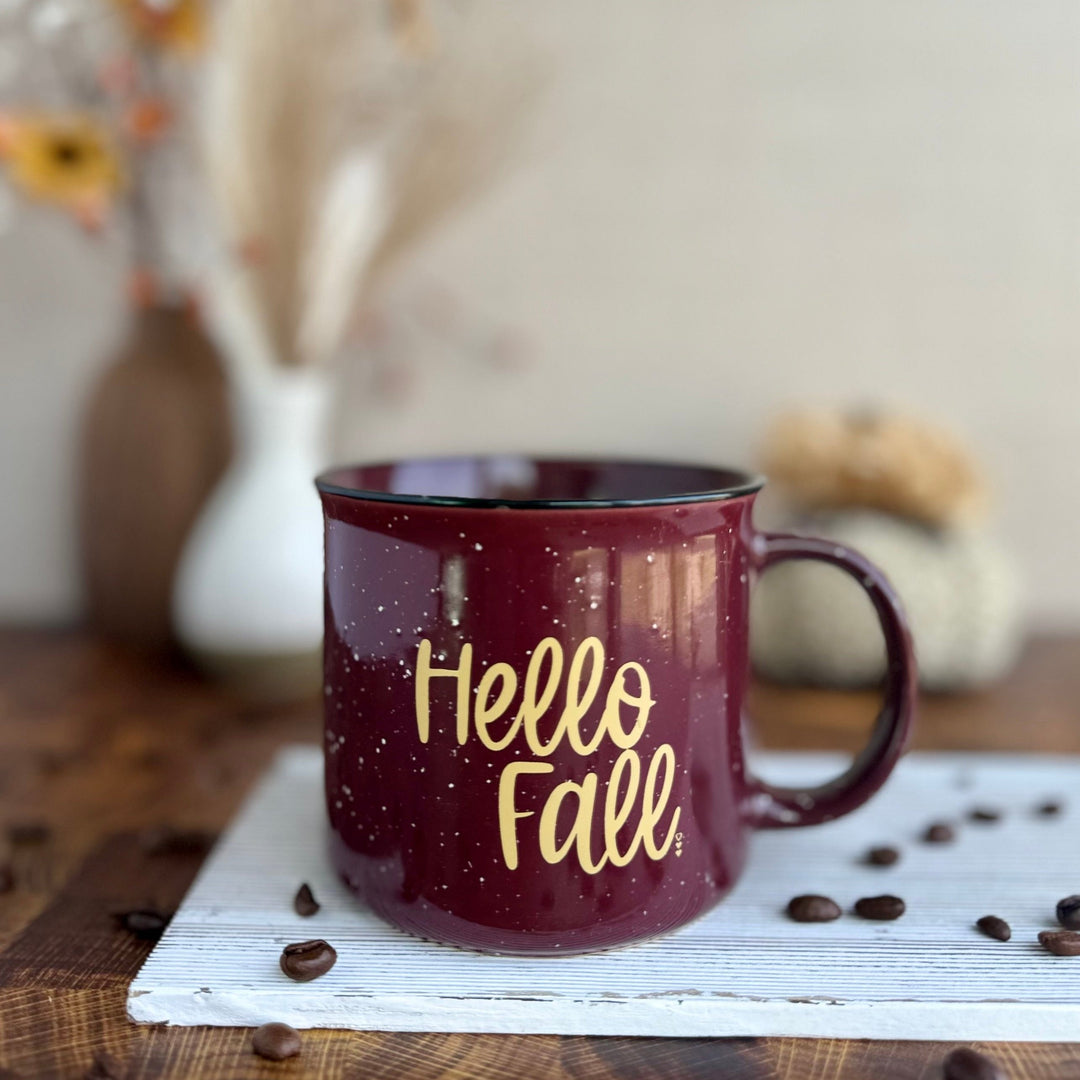 A maroon ceramic coffee mug with black speckles and the text 'Hello Fall' printed in gold on the front, placed on a wooden surface with coffee beans scattered around.