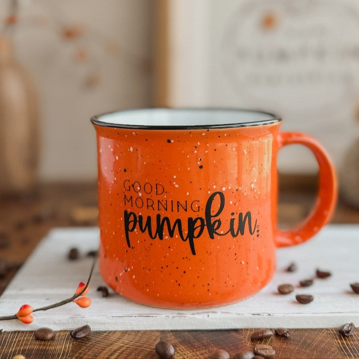 An orange ceramic coffee mug with black speckles and the text 'Good Morning Pumpkin' printed on it, placed on a table with a pumpkin-themed decoration in the background.