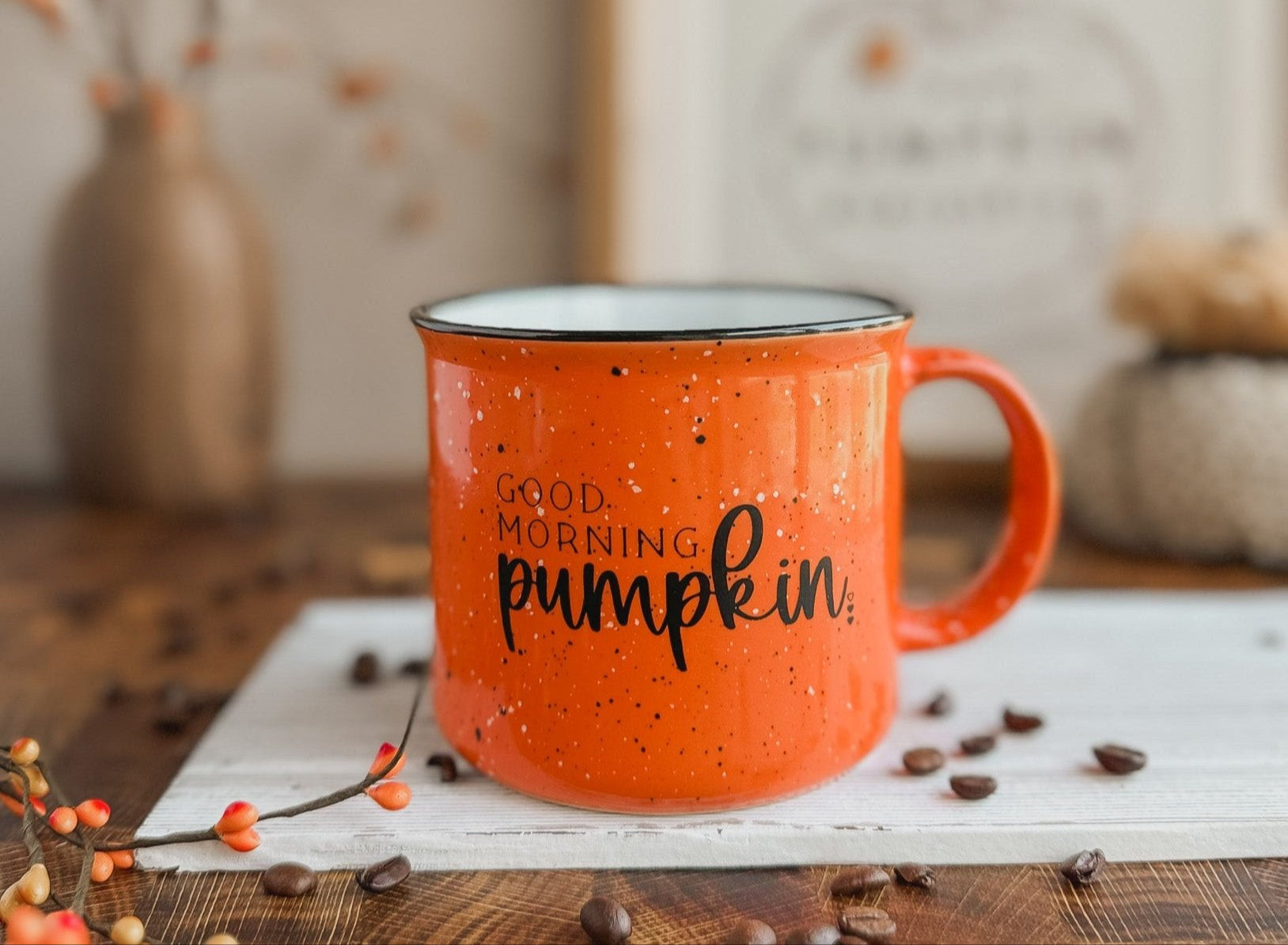 An orange ceramic coffee mug with black speckles and the text 'Good Morning Pumpkin' printed on it, placed on a table with a pumpkin-themed decoration in the background.
