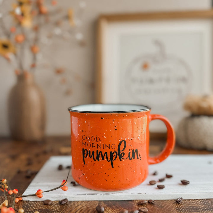 An orange ceramic coffee mug with black speckles and the text 'Good Morning Pumpkin' printed on it, placed on a table with a pumpkin-themed decoration in the background.