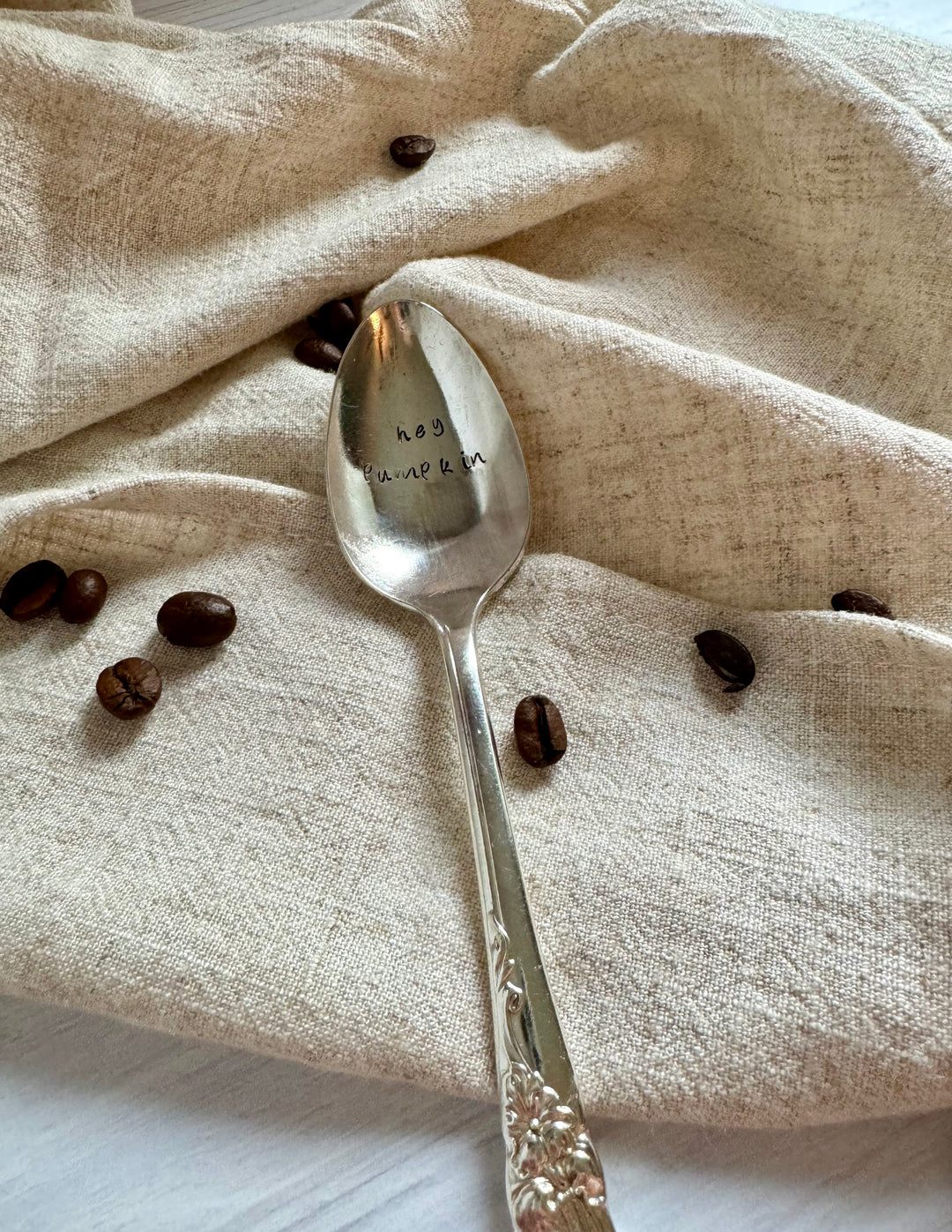 A hand-stamped vintage coffee spoon with the text 'hey pumpkin' on the handle, placed on a beige fabric surface with coffee beans scattered around.