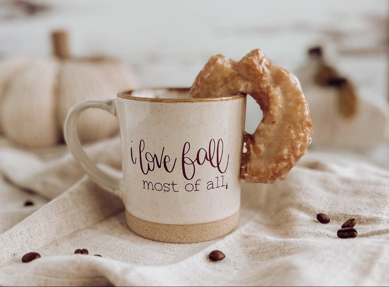 A cream-colored ceramic rustic coffee mug with brown speckles and a maroon text imprint that reads 'I love fall most of all' is shown with a donut in the mug, placed on a linen surface with pumpkins and coffee beans around.