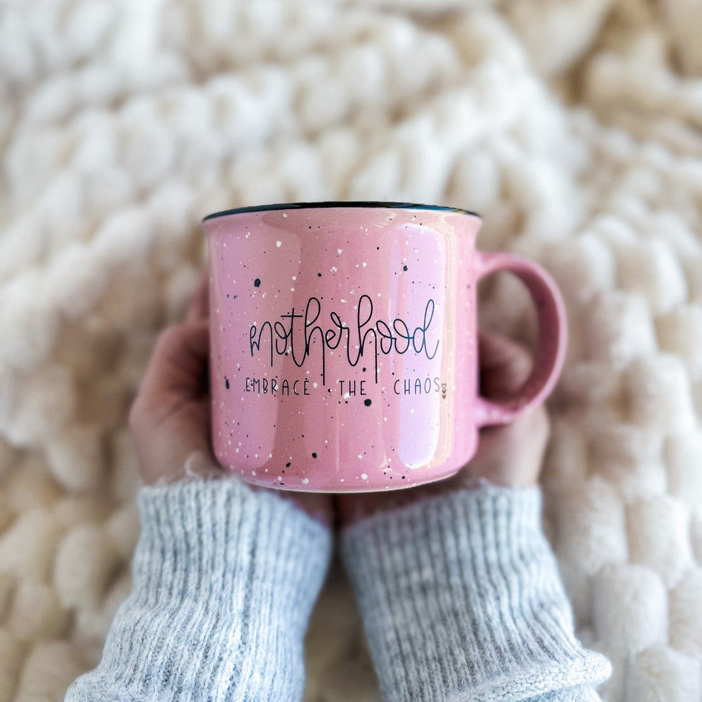 Pink mug with 'motherhood embraces the chaos' text held by a person wearing gray sleeves against a textured beige background.