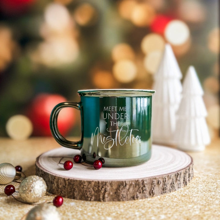 A pearlescent green ceramic coffee mug with the text 'Meet Me Under The Mistletoe' printed in white, placed on a wooden stand surrounded by Christmas decorations including mistletoe, pine branches, and holiday balls.