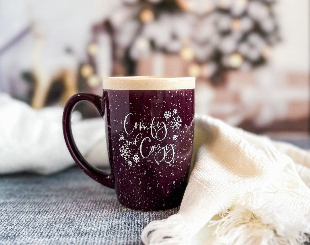 A purple ceramic coffee mug with white speckles and text design, placed on a grey surface next to a white blanket and a Christmas tree in the background.