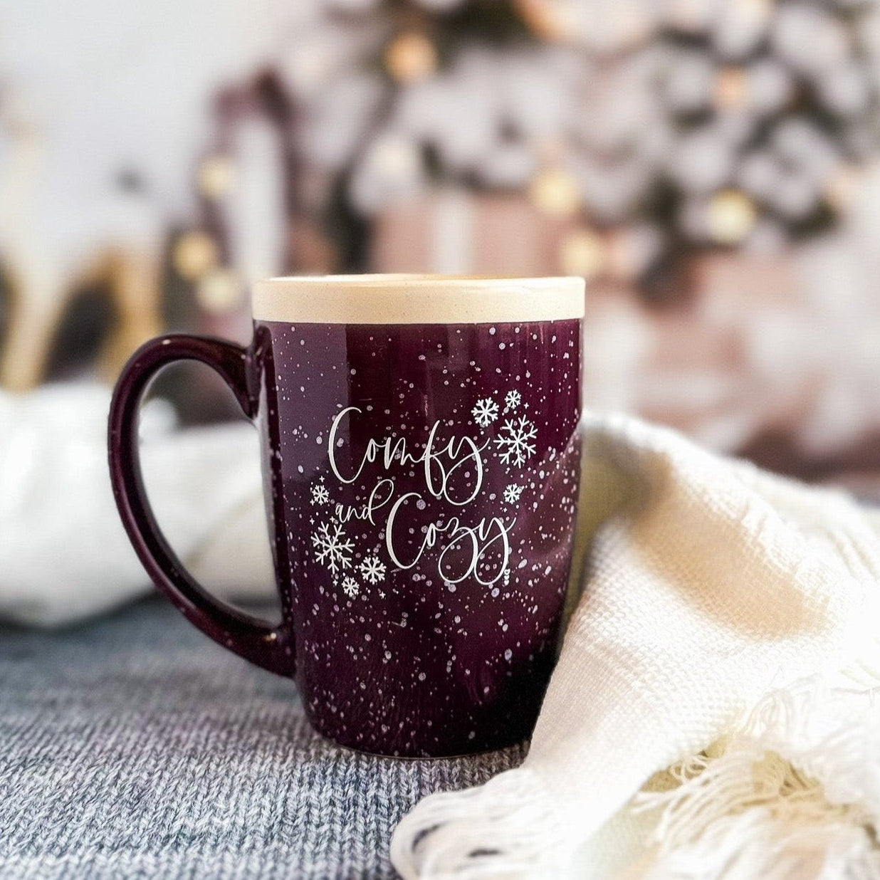 A purple ceramic coffee mug with white speckles and text design, placed on a grey surface next to a white blanket and a Christmas tree in the background.