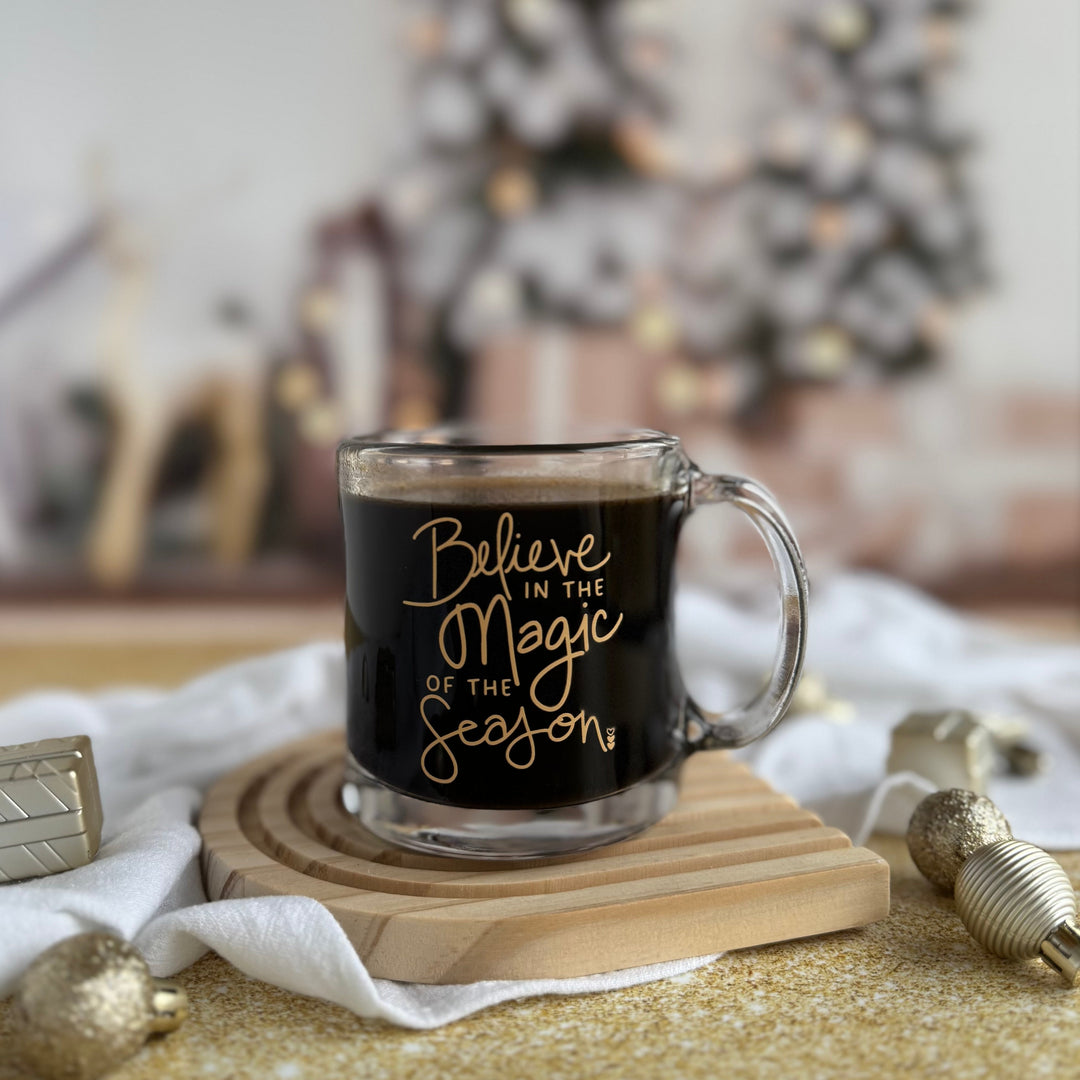 Glass mug with 'Believe in the Magic of the Season' text on a wooden coaster with Christmas decorations in the background.