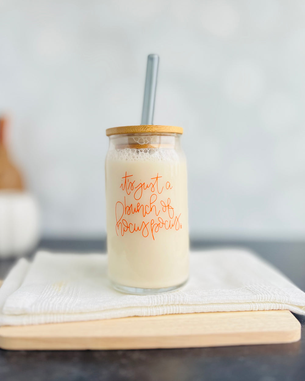 A glass with an orange imprint that reads 'It's Just a Bunch of Hocus Pocus' filled with a beige beverage. A bamboo lid and a straw are also visible.